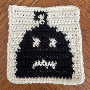 Puede incluir: Posavasos cuadrado de ganchillo hecho a mano con un diseño de fantasma negro sobre un fondo crema. El fantasma tiene dos agujeros para los ojos y una boca. El posavasos está hecho de hilo y tiene un aspecto texturizado.