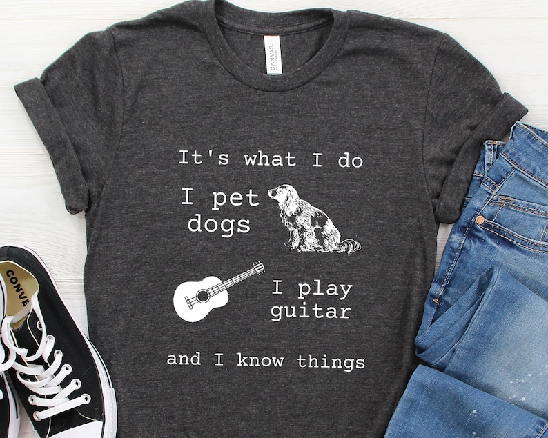 May include: A dark gray t-shirt with white text that reads "It's what I do I pet dogs I play guitar and I know things". The shirt also features a black and white illustration of a dog and a guitar.