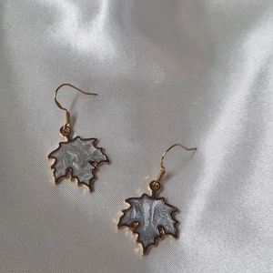May include: A pair of gold-coloured drop earrings. Each earring features a maple leaf design with a white and grey marbled pattern, framed by a gold border. The earrings hang from gold-coloured fish hook ear wires.