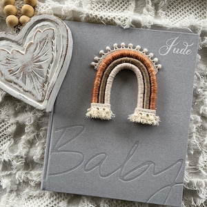 May include: A grey baby book with the word "Baby" embossed on the cover. A small, crocheted rainbow with brown, orange, and cream yarn is on top of the book. The name "Jude" is written in white script on the cover.
