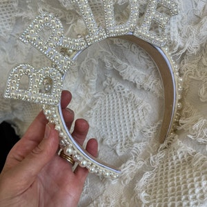 May include: A white headband with the word "Bride" spelled out in pearl-like beads. The headband is made of a flexible material and is decorated with small, white beads.