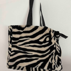 Puede incluir: Una bolsa de mano de estampado de cebra en blanco y negro con asas negras.