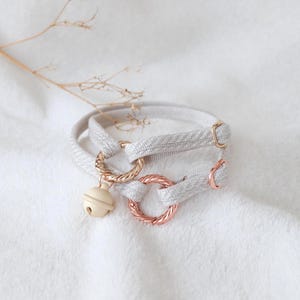 May include: A light gray woven fabric collar featuring two rose gold-toned twisted metal rings and a small, smooth, cream-colored bell charm. The collar includes rose gold-toned metal hardware for adjustment, creating a functional and simple design.