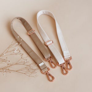 Op de afbeelding: Twee verstelbare banden voor tassen in beige en wit met roségoudkleurige hardware en het woord "Larkspur" gedrukt op elke band.