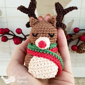 Peut inclure: Une d&eacute;coration de renne en crochet avec un corps brun, un visage blanc, un nez rouge et une &eacute;charpe verte et rouge. Le renne est tenu dans une main.