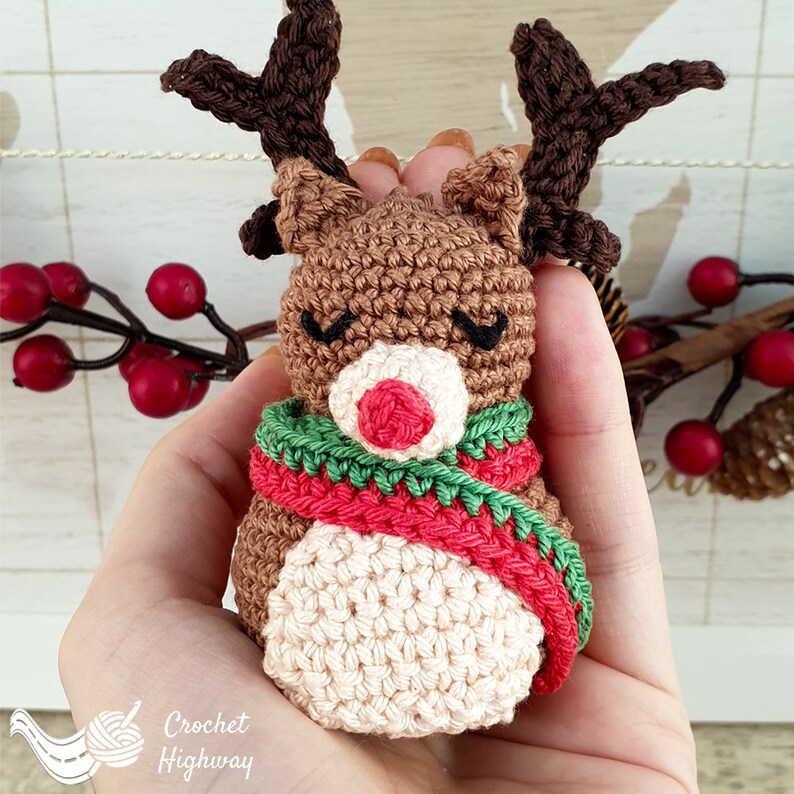 Peut inclure: Une d&eacute;coration de renne en crochet avec un corps brun, un visage blanc, un nez rouge et une &eacute;charpe verte et rouge. Le renne est tenu dans une main.
