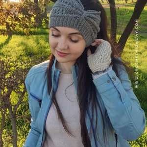 May include: A gray crocheted headband with a knot on the top. The headband is worn by a person with long brown hair. The person is wearing a light blue jacket and a light pink sweater.