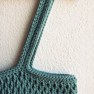 Puede incluir: Una bolsa de mano de crochet de color turquesa con una correa larga colgando de una percha de madera. La bolsa tiene un patr&oacute;n de puntada texturizado y la correa tiene un patr&oacute;n de puntada de espiga. El texto "CROCHETHIGHWAY.COM" es visible en la parte inferior de la imagen.