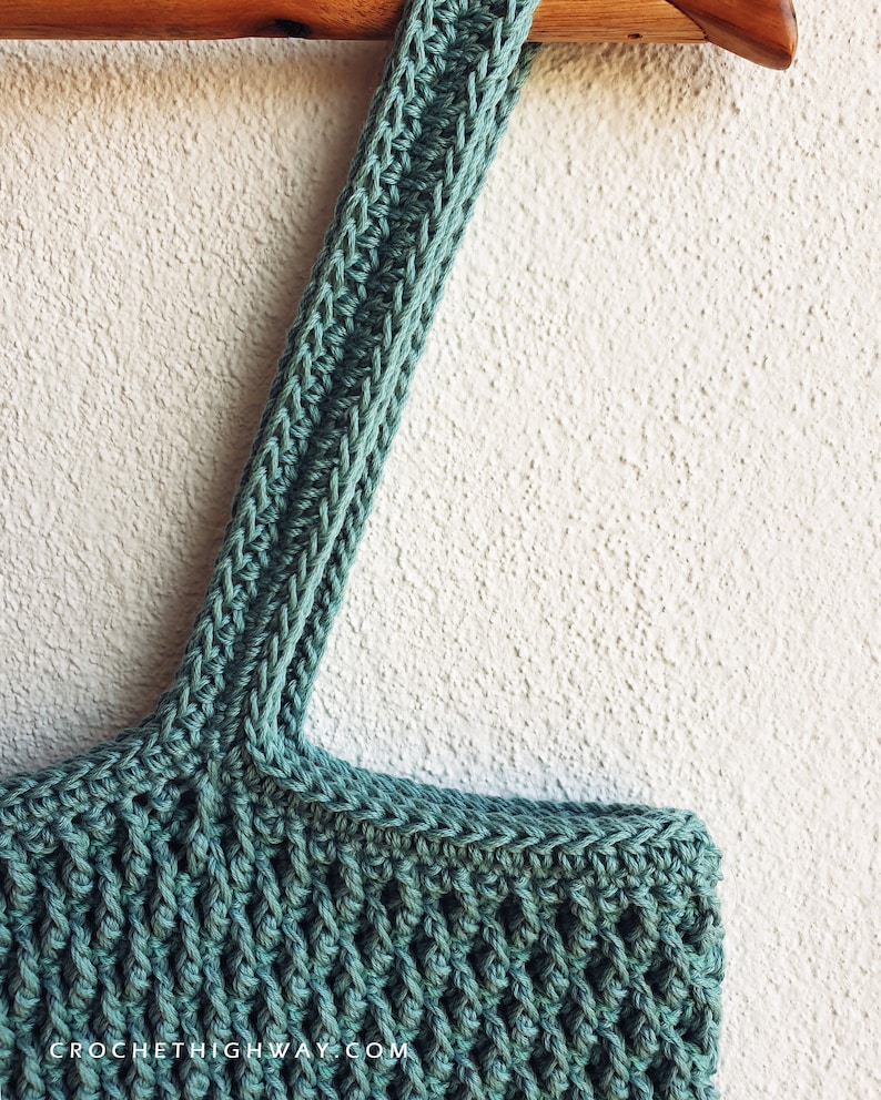 Puede incluir: Una bolsa de mano de crochet de color turquesa con una correa larga colgando de una percha de madera. La bolsa tiene un patr&oacute;n de puntada texturizado y la correa tiene un patr&oacute;n de puntada de espiga. El texto "CROCHETHIGHWAY.COM" es visible en la parte inferior de la imagen.