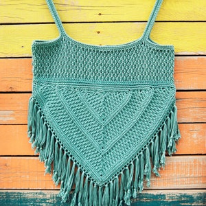 Puede incluir: Un top de crochet de color turquesa con un patr&oacute;n geom&eacute;trico y flecos. El top est&aacute; colgado en una percha de madera sobre un fondo de madera de colores.