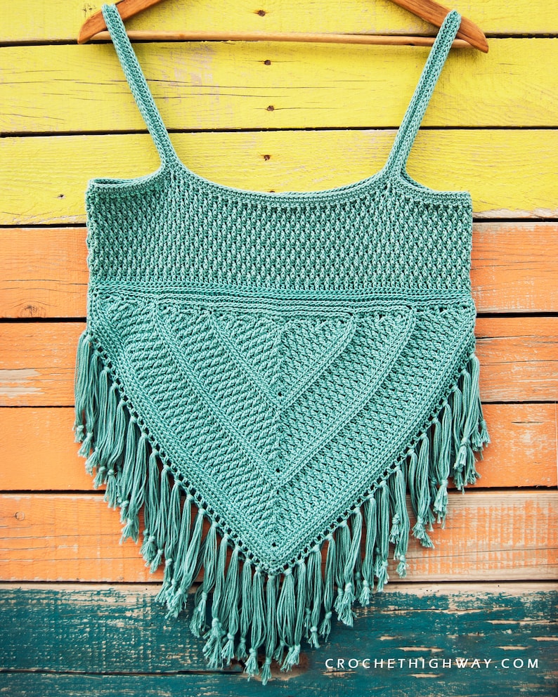 Puede incluir: Un top de crochet de color turquesa con un patr&oacute;n geom&eacute;trico y flecos. El top est&aacute; colgado en una percha de madera sobre un fondo de madera de colores.