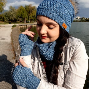May include: A blue crocheted hat, scarf and fingerless gloves set. The hat has a textured stitch pattern and a pompom on top. The scarf and gloves have a similar stitch pattern.