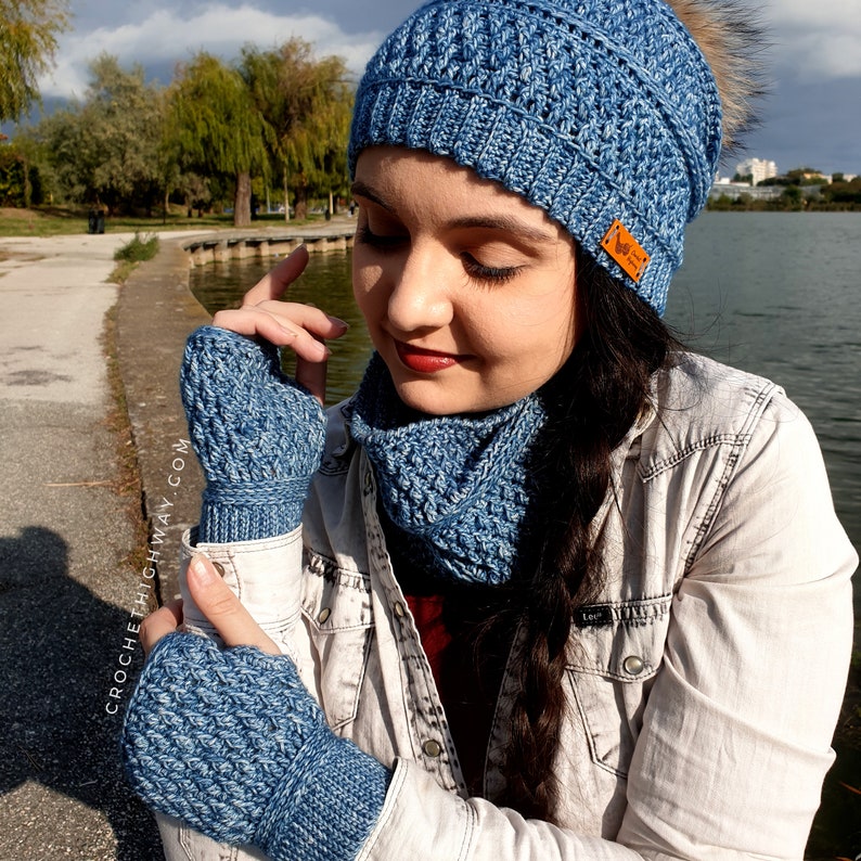 May include: A blue crocheted hat, scarf and fingerless gloves set. The hat has a textured stitch pattern and a pompom on top. The scarf and gloves have a similar stitch pattern.