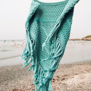 Puede incluir: Un top de crochet de color turquesa con detalles de flecos colgado sobre un fondo de playa. El top tiene un corte holgado y fluido, perfecto para el verano.