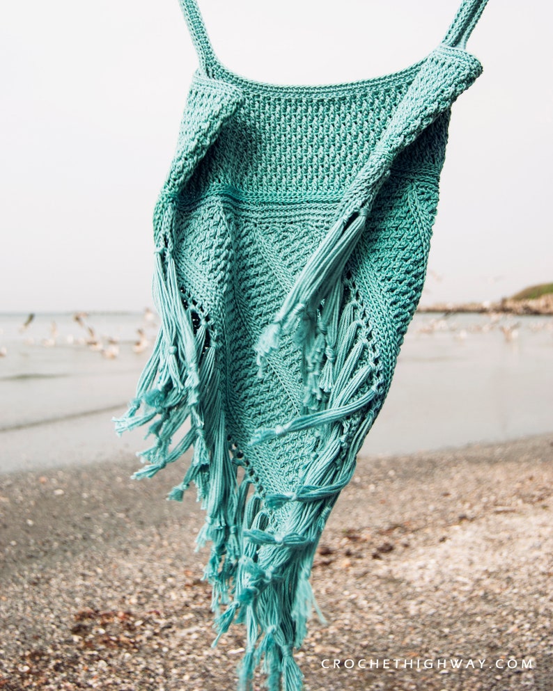 Puede incluir: Un top de crochet de color turquesa con detalles de flecos colgado sobre un fondo de playa. El top tiene un corte holgado y fluido, perfecto para el verano.
