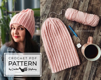 Può includere: Un cappello a uncinetto rosa con una trama a coste. Il cappello è appoggiato su una superficie di legno con un gomitolo di lana rosa, un uncinetto e una tazza di caffè.