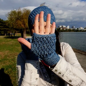 May include: A person wearing a blue crocheted hat and matching fingerless gloves. The person is standing outdoors with a body of water in the background.