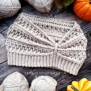 May include: A beige crocheted headband with a criss-cross pattern. The headband is on a wooden surface with balls of yarn and pumpkins.
