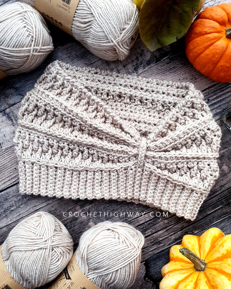 May include: A beige crocheted headband with a criss-cross pattern. The headband is on a wooden surface with balls of yarn and pumpkins.