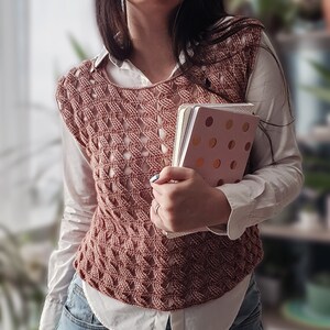K&ouml;nnte beinhalten: Eine rosa geh&auml;kelte Weste mit einem wei&szlig;en Hemd darunter und blauen Jeans. Die Weste hat ein Spitzenmuster und wird von einer Person getragen, die zwei Notizb&uuml;cher h&auml;lt.