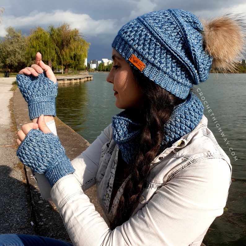 May include: A blue crocheted hat, scarf and fingerless gloves set. The hat has a pompom on top and the scarf is a cowl style. The set is made with a textured stitch.