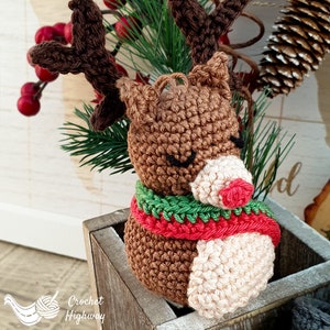 Peut inclure: Une d&eacute;coration de renne en crochet avec une fourrure brune, un nez rouge et une &eacute;charpe verte et rouge. La d&eacute;coration est pos&eacute;e dans une bo&icirc;te en bois.