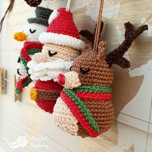 Peut inclure: Trois d&eacute;corations de No&euml;l en crochet&nbsp;: un bonhomme de neige avec un chapeau haut de forme noir, un P&egrave;re No&euml;l avec une barbe blanche et un chapeau rouge, et un renne avec des bois bruns et une &eacute;charpe rouge et verte. Les d&eacute;corations sont suspendues &agrave; un fond en bois.