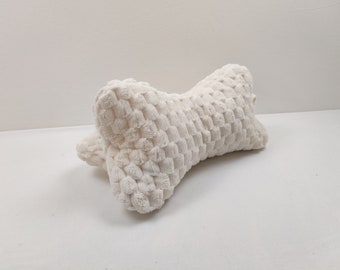 Puede incluir: Una almohada blanca con forma de hueso con una superficie texturizada. La almohada está hecha de tela suave y felpa.