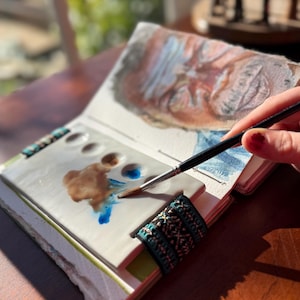 Puede incluir: Cuaderno de bocetos abierto con una pintura de acuarela en progreso. Una paleta de cerámica con pintura marrón y azul está en el centro. Un pincel aplica pintura azul. El cuaderno tiene una banda elástica con estampado turquesa y blanco.