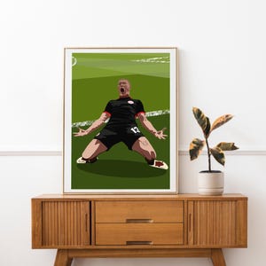 Op de afbeelding: Een ingelijste kunstafdruk toont een voetballer die een doelpunt viert. De speler, in een zwart tenue met rode details en nummer 13, knielt op een groen veld met uitgestrekte armen. Het kunstwerk wordt tentoongesteld op een houten kast.
