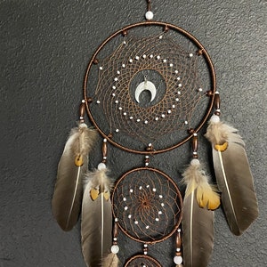 May include: A handcrafted dreamcatcher with a brown circular frame and intricate web design. The dreamcatcher features white beads, brown feathers, and a white crescent moon charm in the center. The design is suspended against a dark gray textured background.