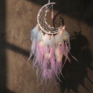 May include: A crescent moon dreamcatcher with a white web, adorned with pink and white feathers. The dreamcatcher has small lights and a pink crystal pendant hanging from it. The background is a textured, brown wall.