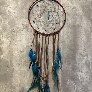 May include: A brown dreamcatcher with a circular woven web design. The dreamcatcher features turquoise beads, blue and teal feathers, and a turquoise pendant in the center. The dreamcatcher is suspended by a brown cord.