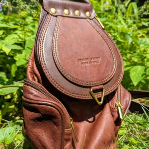 Peut inclure: Un sac à dos en cuir marron avec des ferrures dorées et une fermeture à rabat. Le sac à dos a une poche zippée à l'avant et une étiquette qui indique "Cuir véritable Fabriqué en Italie".