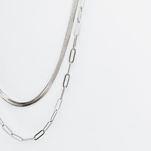 May include: Two silver chains, one a snake chain and the other a chain with oval links, are layered on a white background.