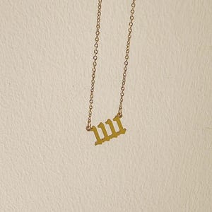 Puede incluir: Un collar dorado con una cadena y un colgante con el n&uacute;mero 1111 en una fuente inglesa antigua. El collar cuelga sobre un fondo neutro.