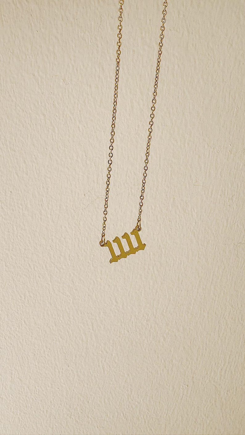 Puede incluir: Un collar dorado con una cadena y un colgante con el n&uacute;mero 1111 en una fuente inglesa antigua. El collar cuelga sobre un fondo neutro.