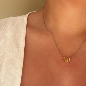 Puede incluir: Un collar dorado con un colgante con el n&uacute;mero 1111. El collar se lleva sobre una parte superior blanca. El fondo es de color beige neutro.