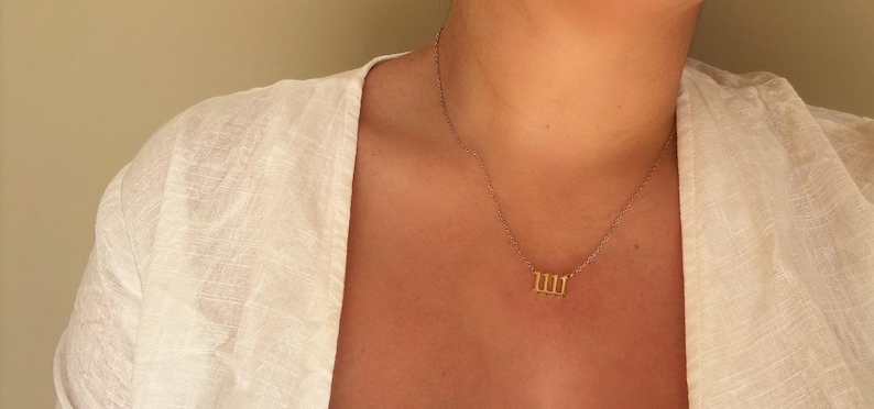 Puede incluir: Un collar dorado con un colgante con el n&uacute;mero 1111. El collar se lleva sobre una parte superior blanca. El fondo es de color beige neutro.