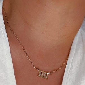 Puede incluir: Un collar dorado con una cadena delicada y un colgante con el n&uacute;mero romano "III". El collar se muestra sobre un fondo de tela blanca.