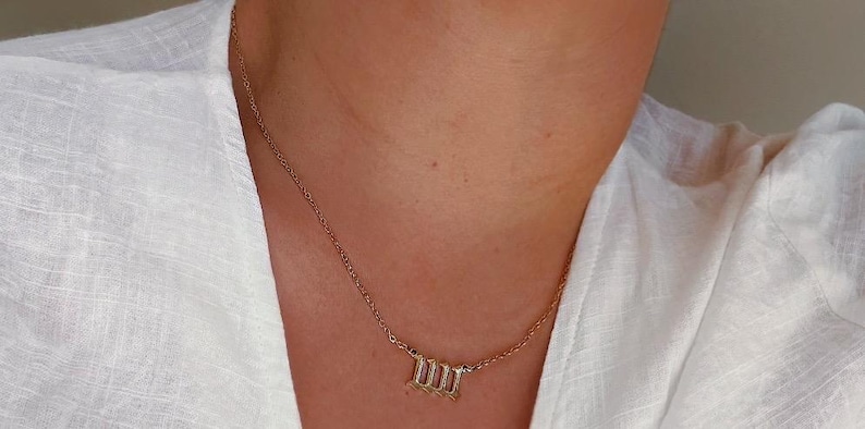 Puede incluir: Un collar dorado con una cadena delicada y un colgante con el n&uacute;mero romano "III". El collar se muestra sobre un fondo de tela blanca.