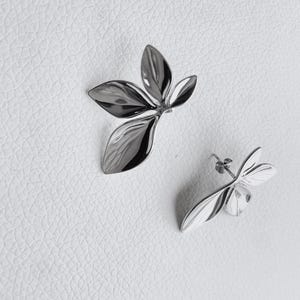 May include: A pair of silver-coloured leaf-shaped earrings and a matching brooch. The earrings have a post-back closure. The brooch and earrings are made of a shiny, reflective metal. The jewellery is set against a white textured background.