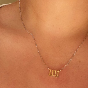 Puede incluir: Un collar dorado con un colgante con el n&uacute;mero 111. El collar lo lleva una persona que viste una parte superior blanca. La cadena es delicada y el colgante es un numeral estilizado.