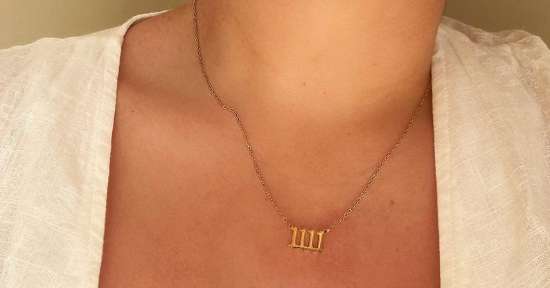 Puede incluir: Un collar dorado con un colgante con el n&uacute;mero 111. El collar lo lleva una persona que viste una parte superior blanca. La cadena es delicada y el colgante es un numeral estilizado.