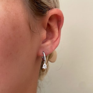May include: A silver hoop earring with a teardrop-shaped diamond dangling from it. The earring is set with small diamonds around the hoop.