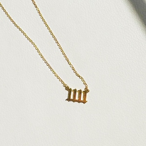 Puede incluir: Collar dorado con un colgante que muestra el n&uacute;mero 111 en una fuente inglesa antigua. La cadena es delicada y el colgante est&aacute; centrado. El collar est&aacute; sobre una superficie blanca.
