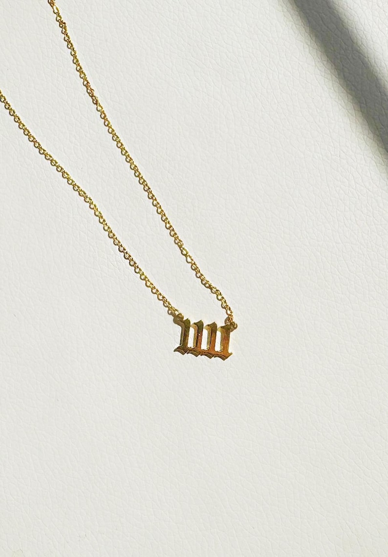 Puede incluir: Collar dorado con un colgante que muestra el n&uacute;mero 111 en una fuente inglesa antigua. La cadena es delicada y el colgante est&aacute; centrado. El collar est&aacute; sobre una superficie blanca.