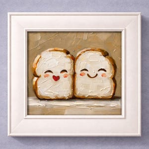 May include: A framed painting featuring two slices of bread with smiling faces and rosy cheeks. The bread has a white, textured topping and a brown crust. One slice has a red heart, and the other has a curved smile. The painting is in a white frame.