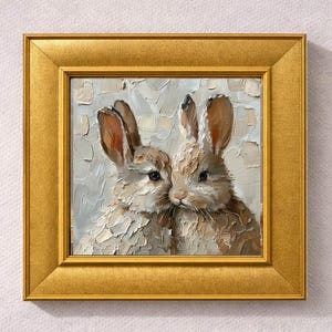 May include: A framed oil painting featuring two rabbits with textured fur and long ears. The rabbits are painted in shades of brown and white, set against a light gray background. The artwork is enclosed in a gold-colored frame.