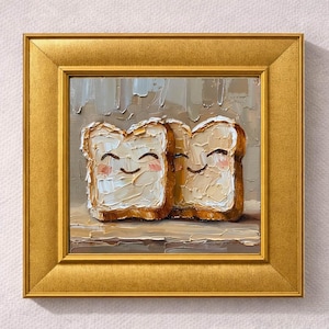 May include: A framed oil painting depicts two slices of toast with smiling faces. The toast has a golden-brown crust and a creamy, textured filling. The artwork is set against a neutral background and framed in a gold-colored frame.